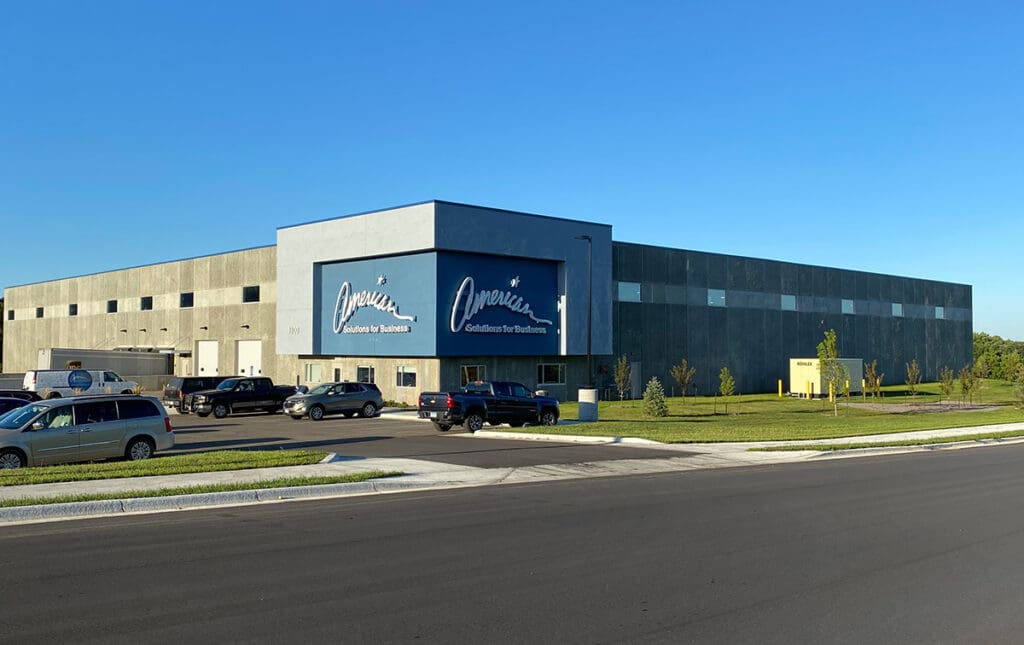 A large, gray industrial warehouse with the "American Solutions for Business" logo on its facade, several cars parked in front, grass and young trees nearby, all beneath a clear blue sky.