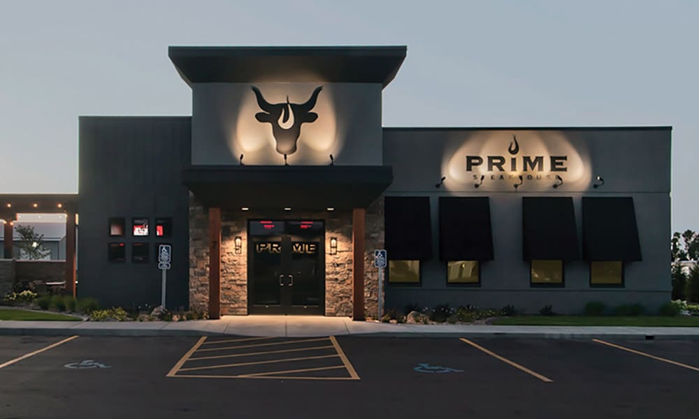 A modern steakhouse restaurant with a bull logo above the entrance and the word "PRIME" on the building, this spot is a favorite among city administration workers. The gray exterior features black awnings, stone accents, and a well-lit parking lot in front.
