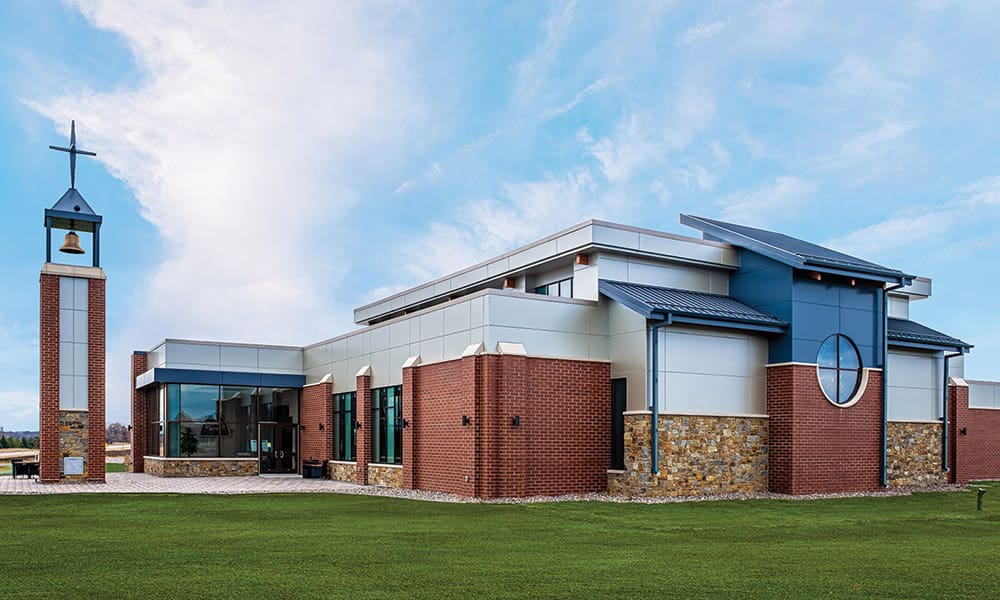 Modern church building with red brick, white paneling, and stone accents stands tall near the municipality center, featuring a bell tower with a cross and circular windows, set against a blue sky with scattered clouds.