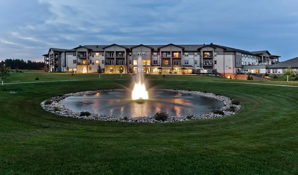 A large, modern building with multiple balconies is seen at dusk, lights glowing from windows. In front of Crystal Brook Senior Living, a circular pond with a bright fountain is surrounded by green, neatly manicured lawns.