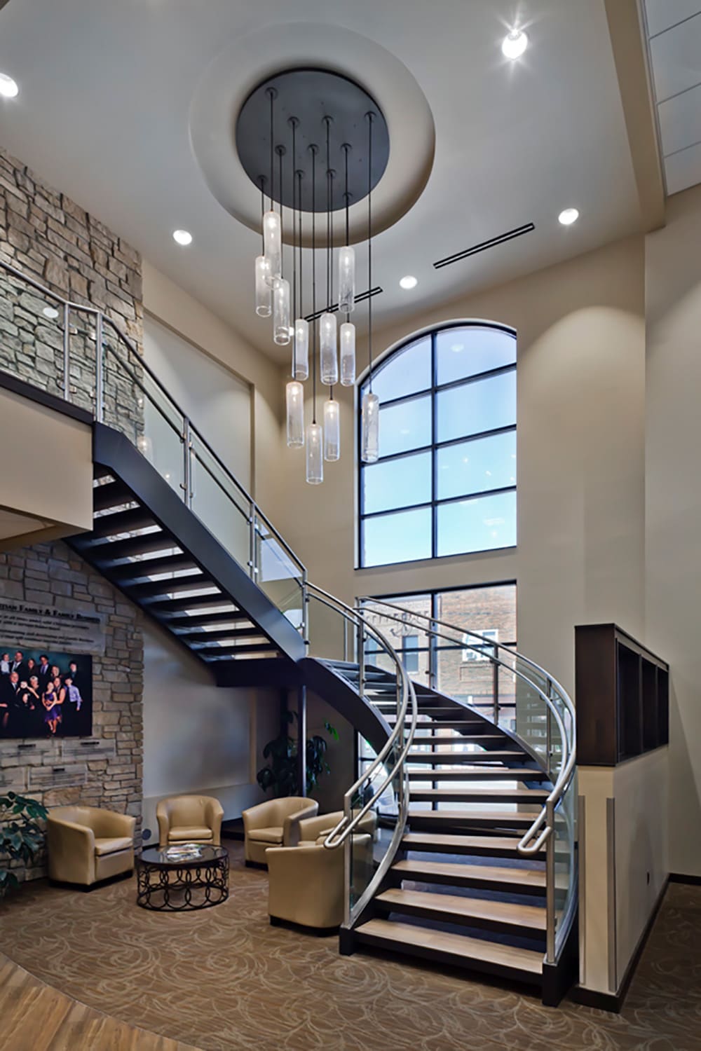 Modern lobby at Glenwood State Bank with a curved staircase, glass railings, beige armchairs around a small table, tall windows, stone accent wall, and a contemporary chandelier hanging from the high ceiling.
