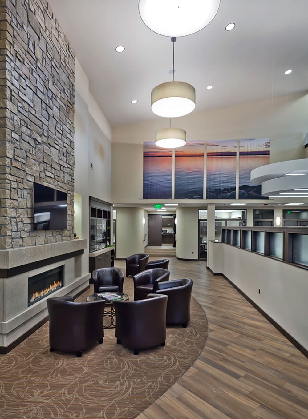 Modern lobby at Glenwood State Bank with a stone fireplace, round ceiling lights, four dark leather chairs around a small table, wood-patterned floor, and a large multi-panel sunset photo on the wall.