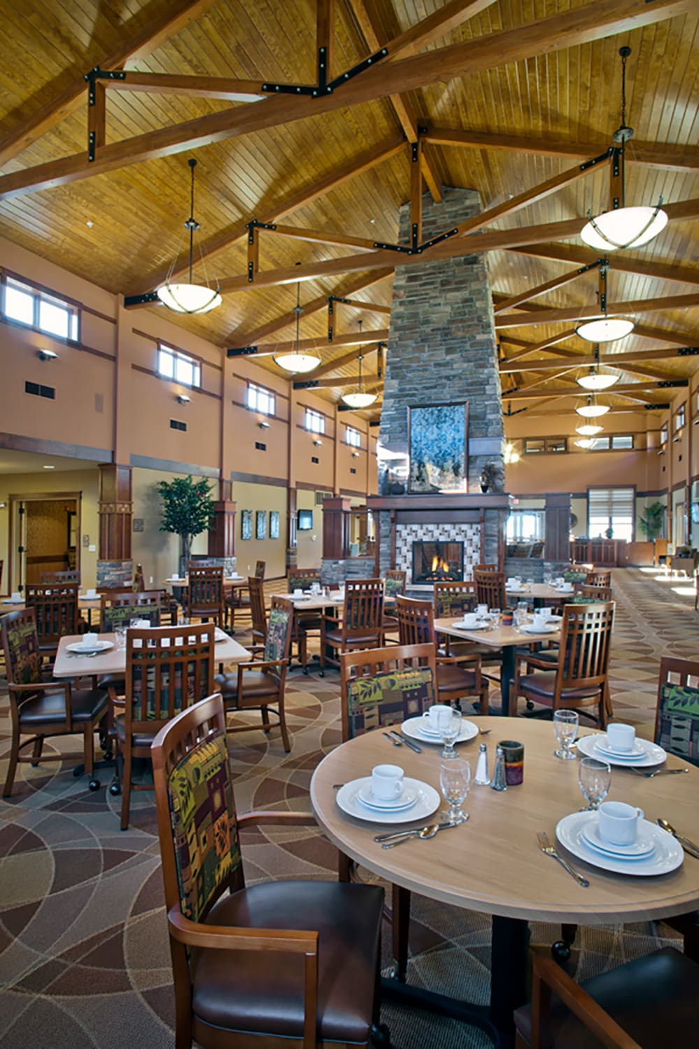 Spacious, elegant restaurant at Grand Arbor by Knute Nelson features wooden ceiling beams, a large stone fireplace, round tables with white dishes and glassware, and neatly arranged wooden chairs—creating a warm, inviting space for senior living residents.