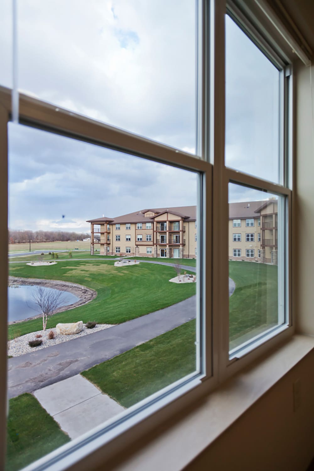 View through a window at Grand Arbor by Knute Nelson reveals a large senior living apartment building, green lawns, a pond with rocks, and a concrete pathway outside under a partly cloudy sky.