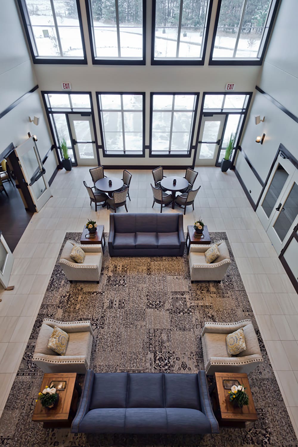 The Crystal Brook Senior Living lobby is spacious and modern, featuring large windows, two dark sofas, patterned armchairs, coffee tables, potted plants, and small dining tables with chairs—all seen from above with snow visible outside.
