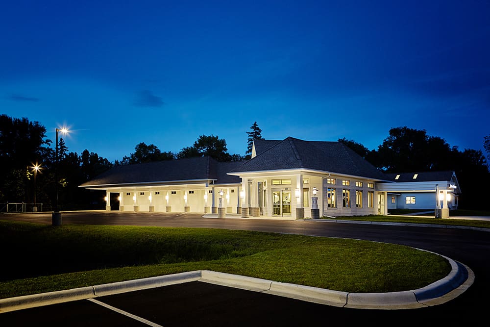 A well-lit, modern building with large windows and columns stands surrounded by trees at dusk in Waite Park. The deep blue sky looms above as the empty parking lot of Executive Express curves around the structure.