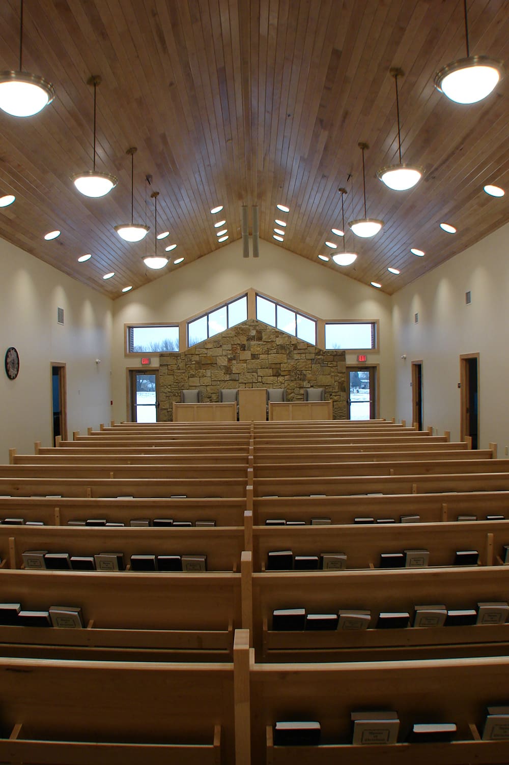 A view of the Morris North Apostolic Church interior with wooden pews, hymnals, and a wooden ceiling. The altar area features a stone wall, several chairs, and large windows filling the Apostolic Church with natural light.