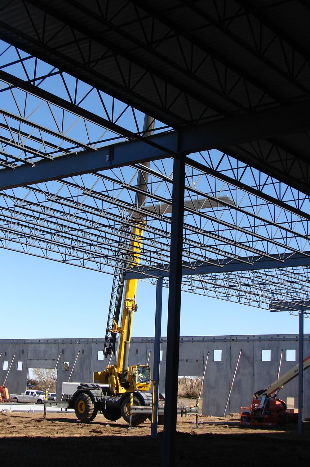 A yellow Clyde Machine crane lifts steel beams at a construction site for a large building, with exposed metal framework and concrete walls. Several vehicles and workers are visible in the background under a clear sky.