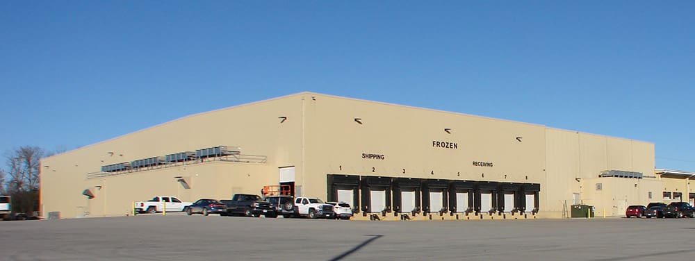 A large beige warehouse belonging to Henry's Foods features multiple loading docks labeled "Frozen," "Shipping," and "Receiving." Several cars and trucks are parked in front under a clear blue sky.