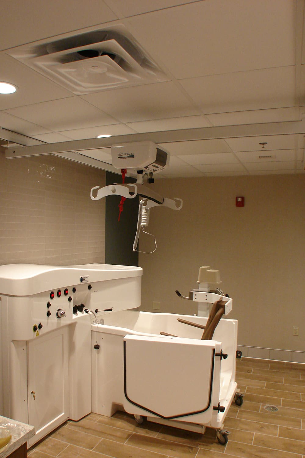 A hospital room at St. Williams Living Center with a specialized white bathtub and adjustable lift system, designed for senior care and assisted living, set against beige tiled walls and wood-look flooring.