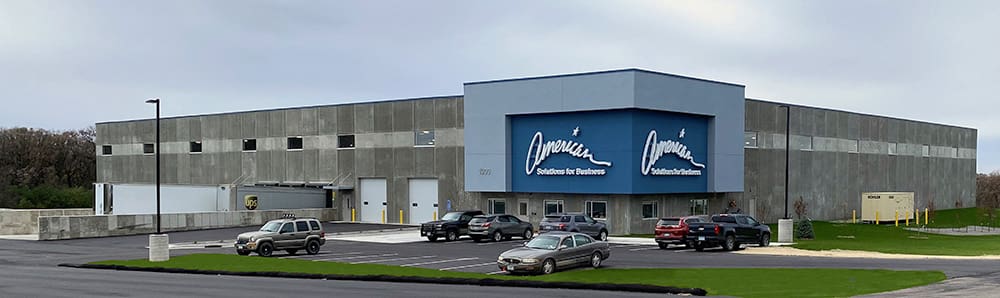 A large, modern warehouse-style building with a gray exterior, several parked cars in front, and two blue signs displaying the words 