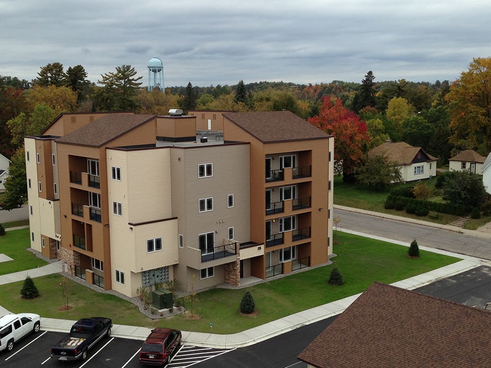 A four-story beige and brown apartment building with balconies sits on a corner lot along 1st Avenue, surrounded by trees with autumn foliage. Several parked cars and a water tower are visible in the background under a cloudy sky.