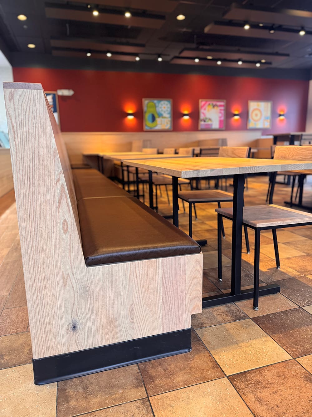 A modern Qdoba restaurant interior with wooden booths, tables, and chairs. The floor has brown tiles, and the red walls feature decorative art and wall-mounted lights. The space appears clean and empty in this Qdoba Refresh setting.
