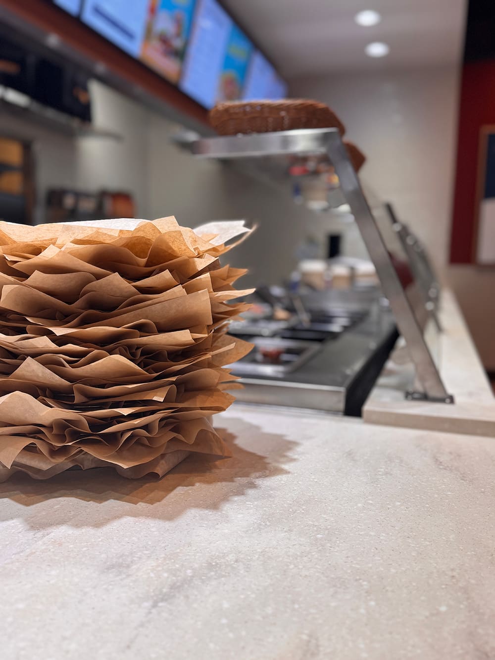 A stack of brown sandwich wraps sits on a counter in front of a deli food preparation area, with various sandwich toppings and digital Qdoba menu boards visible in the background.