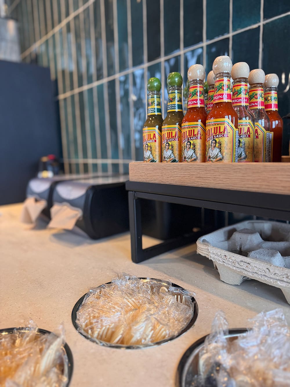 Bottles of Cholula hot sauce are displayed on a counter next to containers of wrapped wooden utensils, adding flavor and spice to the Qdoba menu in this restaurant or café setting.