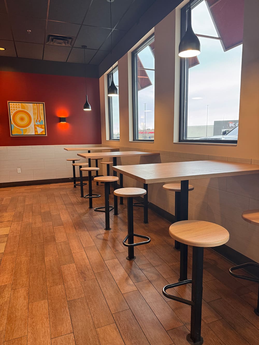 A fast food restaurant interior inspired by the Qdoba Refresh, with round wooden stools, long tables, wood-patterned flooring, red accent walls, modern art, wall light, and large windows letting in natural light.
