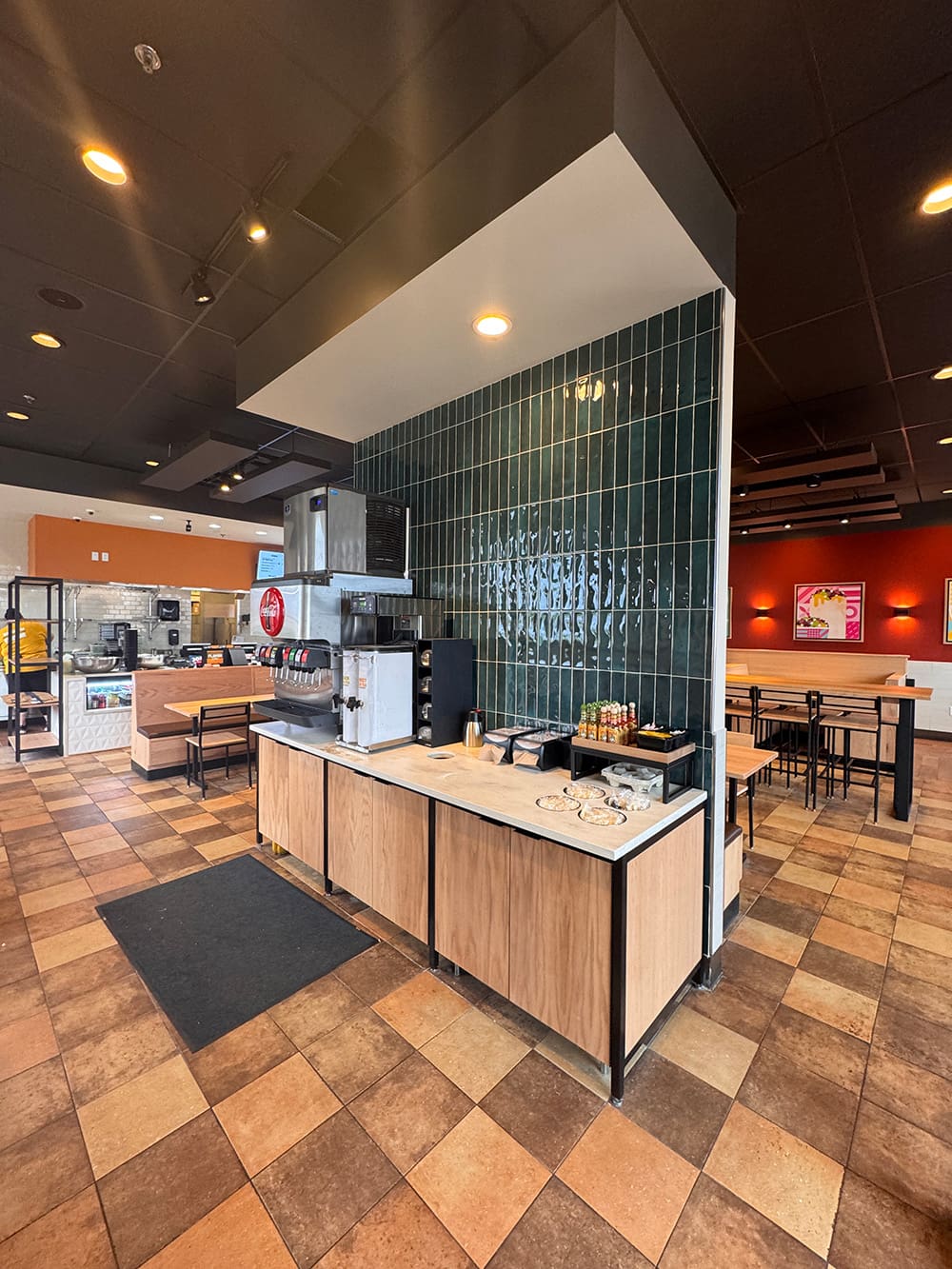 A restaurant interior featuring a self-serve drink and condiment station in the foreground, wood and tile decor, and tables and chairs in the background under warm lighting—just like you’d find at Qdoba while exploring their Qdoba menu.
