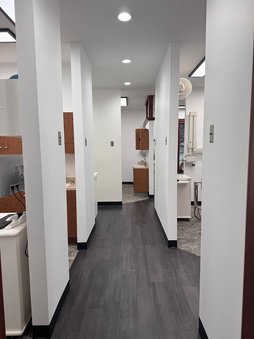 A clean, modern hallway in a dental or medical office with white walls, dark flooring, bright overhead lights, and examination rooms on both sides, featuring cabinetry and dental equipment.