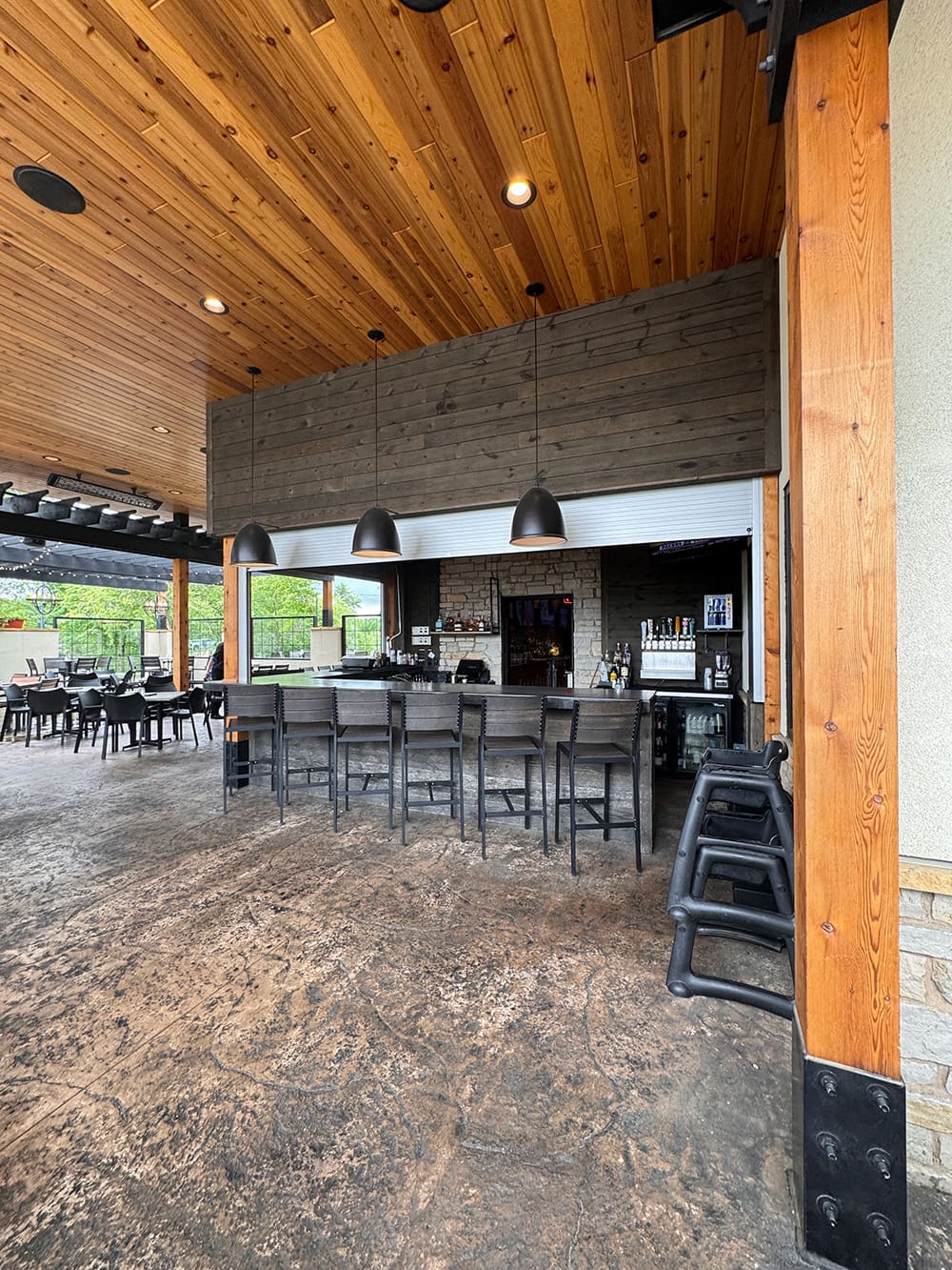 Outdoor bar area with tall wooden ceiling, pendant lights, high chairs at the bar, and empty dining tables. The floor is textured concrete and large windows reveal greenery outside. Stacked chairs are beside a pillar.