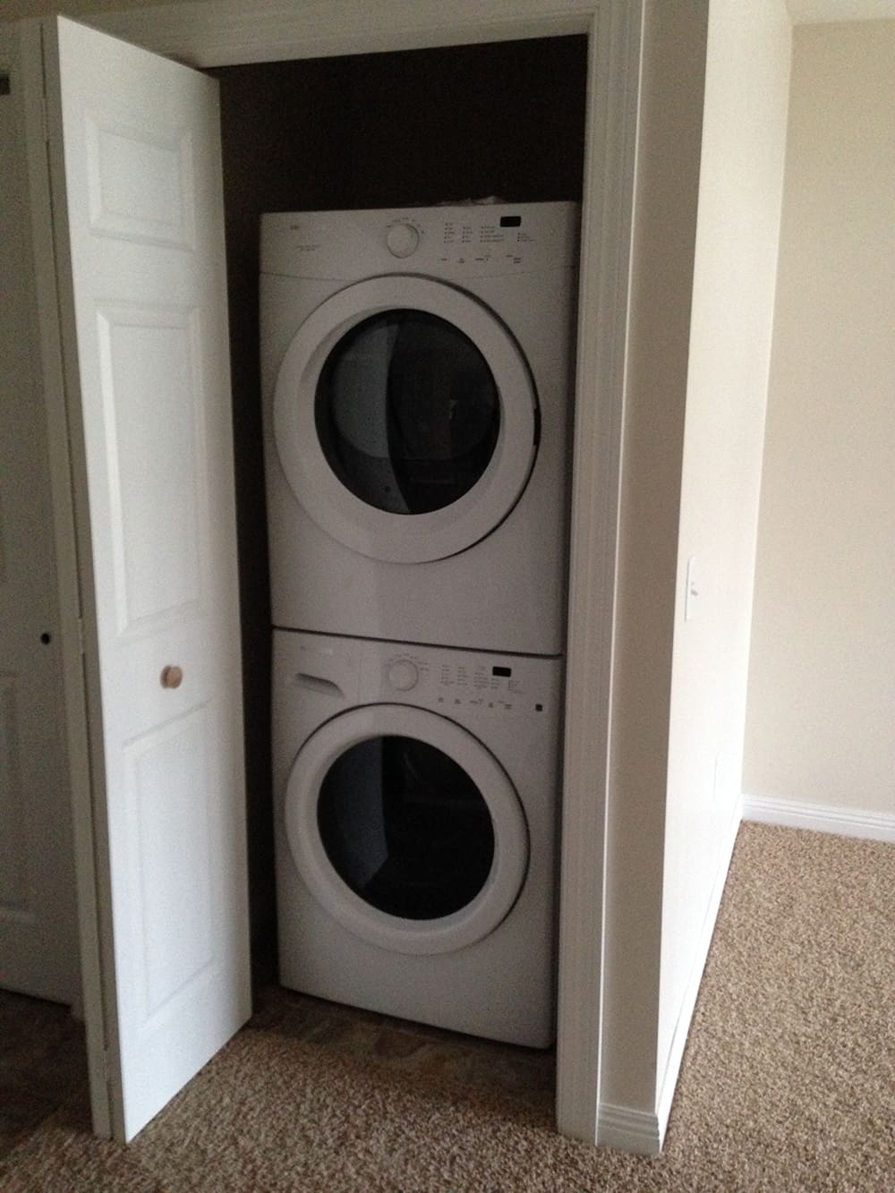A stacked washer and dryer set is placed inside a small closet with the white door partially open, in a carpeted room with beige walls—ideal for convenient living in 1st Avenue rental condos.