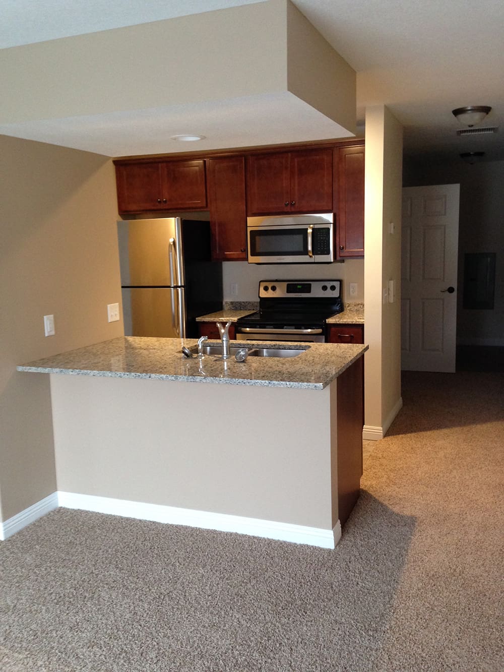A modern apartment kitchen in rental condos on 1st Avenue, featuring stainless steel appliances, dark wood cabinets, granite countertops, a breakfast bar with sink and faucet, and beige carpeted flooring extending into the living area.