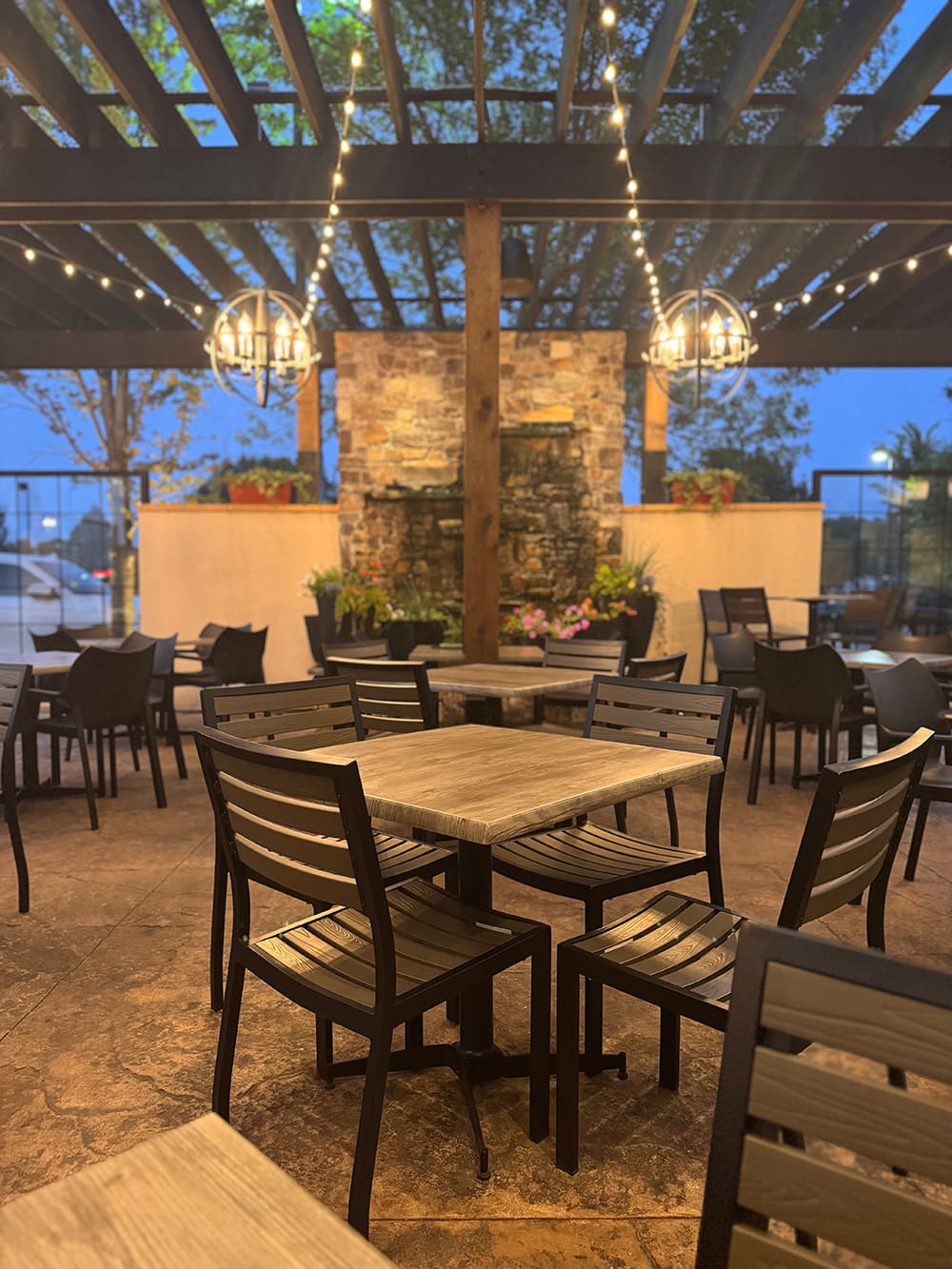 Outdoor patio dining area with wooden tables and black chairs, string lights overhead, stone fireplace in the background, and trees visible through the open pergola roof. Evening ambiance with soft, warm lighting.