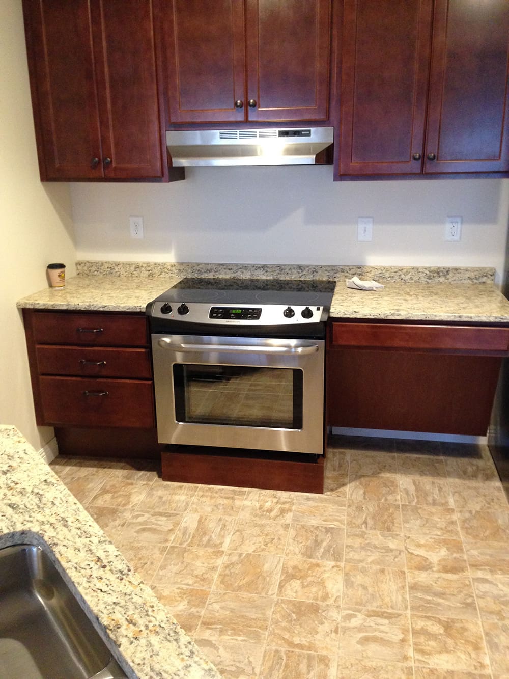A modern kitchen in 1st Avenue rental condos features brown wooden cabinets, granite countertops, a stainless steel electric stove with oven, a range hood, and beige tiled flooring. A cup and a cloth rest on the counter.