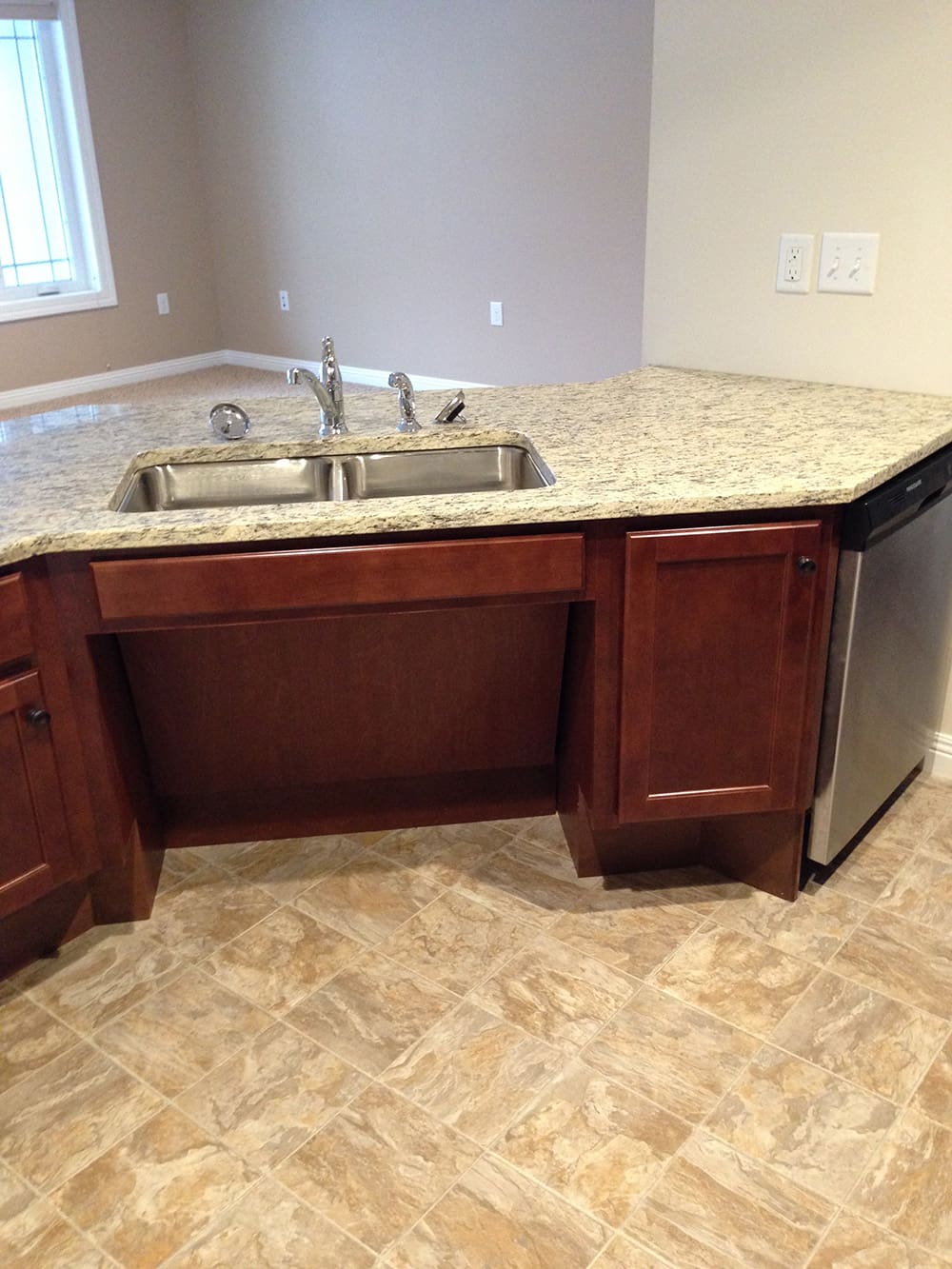 A kitchen area with a granite countertop, double sink with faucet, wooden cabinets, stainless steel dishwasher, and beige tiled floor. Perfect for rental condos on 1st Avenue, the space features light brown walls and a window in the background.
