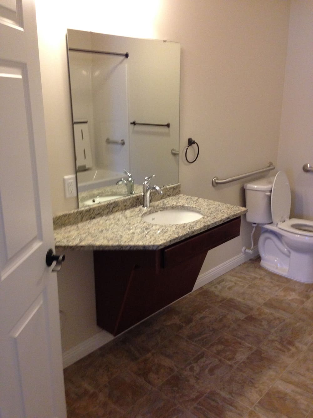 Accessible bathroom in 1st Avenue rental condos featuring a granite countertop sink, large mirror, wall-mounted grab bars near the toilet, and tan tile flooring. The sink is designed for wheelchair accessibility.