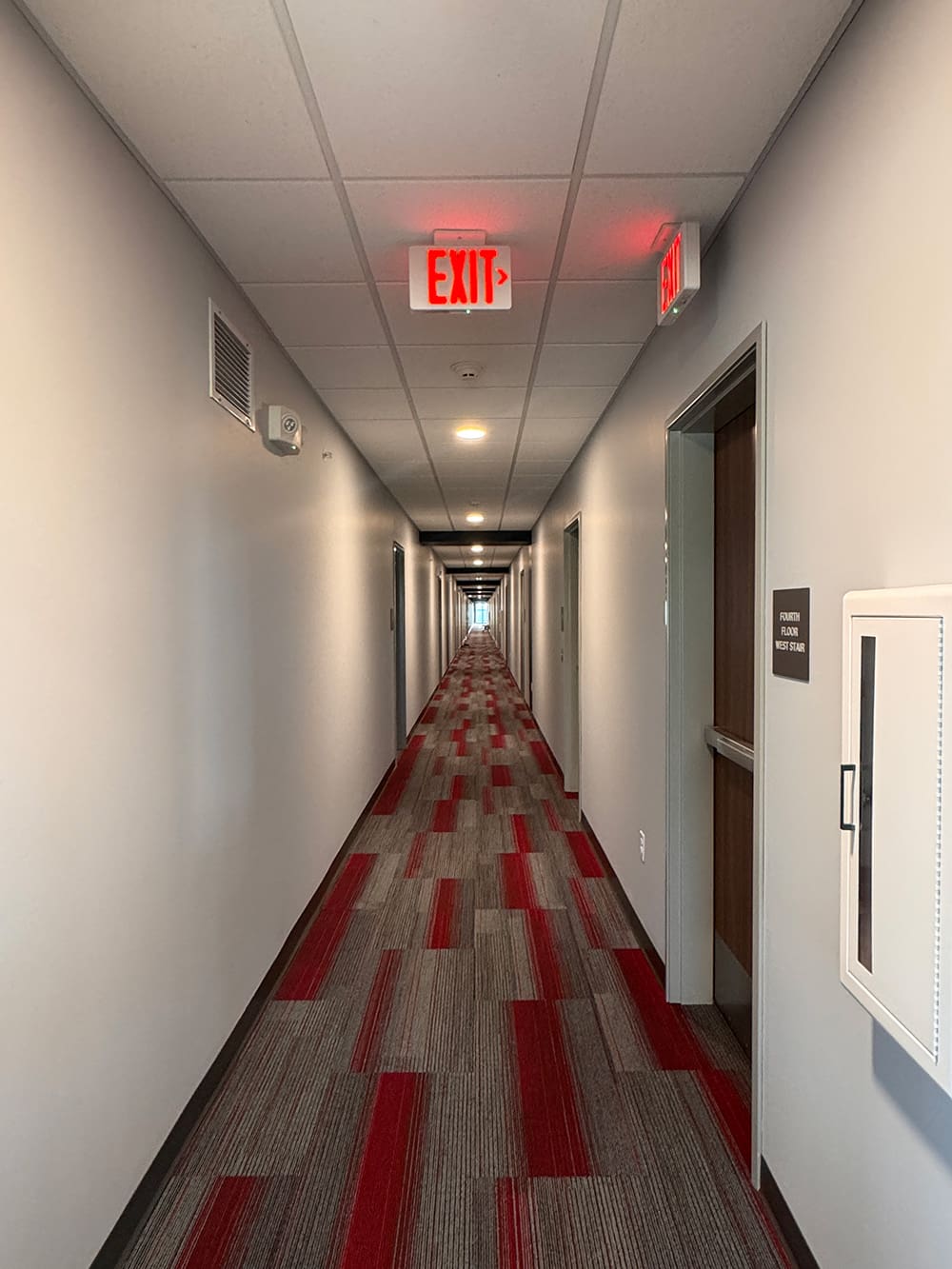 A long, narrow hallway with gray walls and a red-and-gray patterned carpet in ATCC Foundation Hall; doors line both sides, and red EXIT signs are visible on the ceiling at either end, typical of modern student housing.
