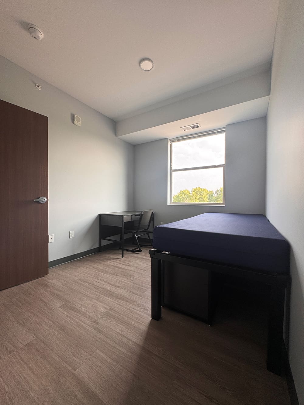 A small, minimalist dorm room in ATCC Foundation Hall with light walls, wooden floor, a closed wooden door, a single bed with a blue mattress, a black desk and chair, and a window letting in natural light—perfect student housing.