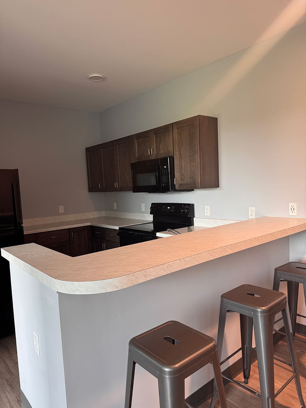 Modern kitchen with a light-colored countertop, dark wood cabinets, and three metal barstools along the breakfast bar—perfect for student housing at ATCC Foundation Hall. The space features black appliances, light gray walls, and a clean, minimalist design.