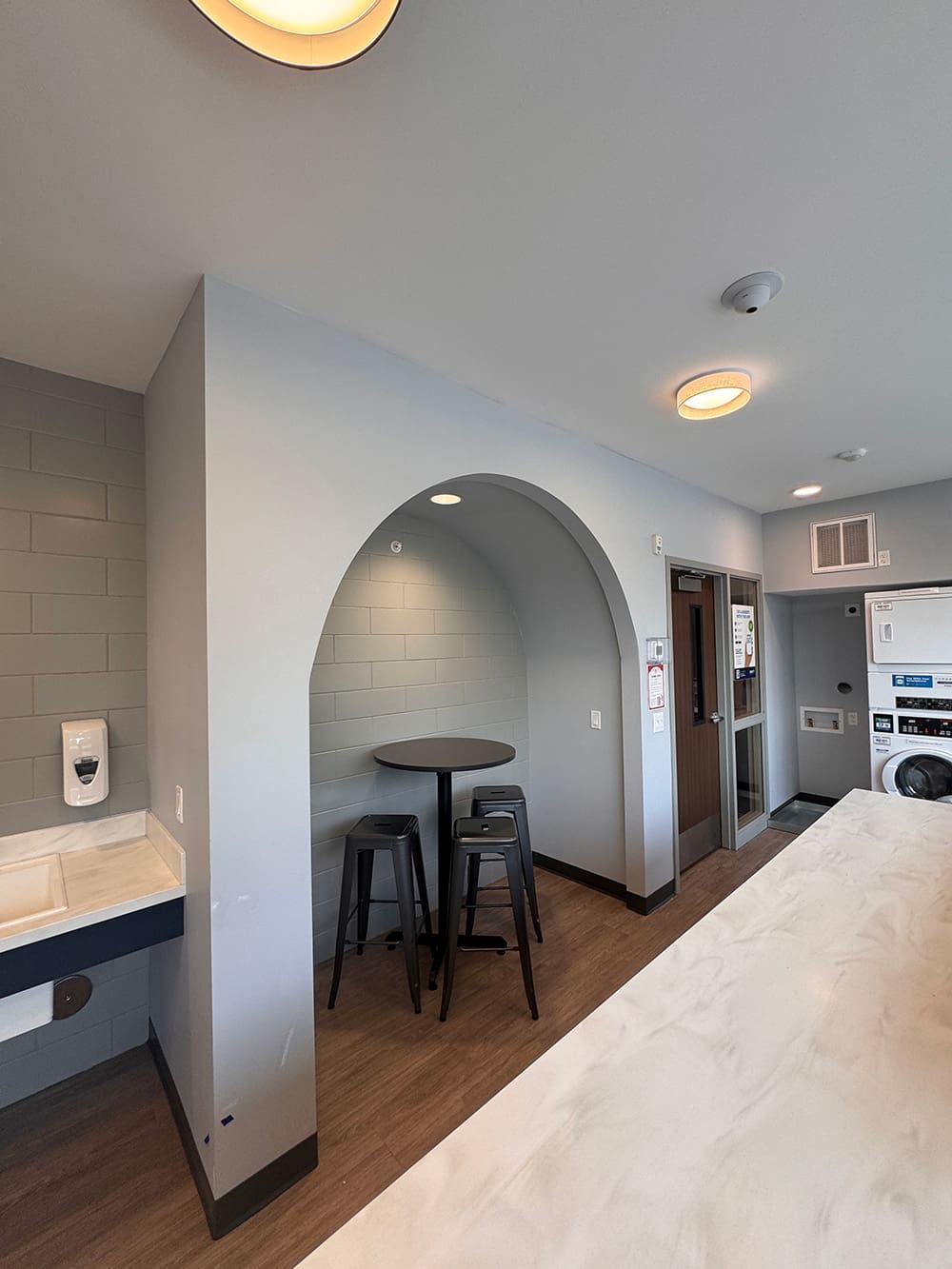 A small seating nook with a round table and two black stools sits under an archway in a modern laundry room with gray walls and wood flooring—perfect for student housing like ATCC Foundation Hall, with a washer and dryer visible in the background.