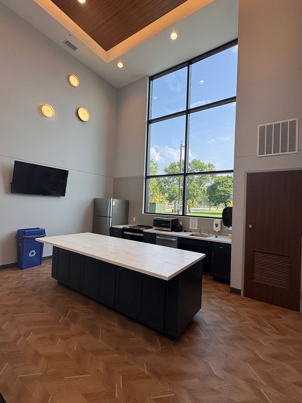 A modern kitchen in student housing with a large island, stainless steel appliances, wall-mounted TV, recycling bin, and tall windows letting in natural light with tree views. The wood-patterned floor adds style to this ATCC Foundation Hall space.