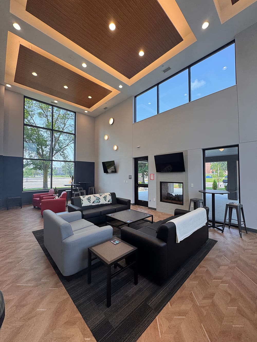 A spacious, modern lounge in ATCC Foundation Hall with tall windows, multiple sofas, armchairs, side tables, a fireplace, wall-mounted TVs, and wood-patterned flooring. Natural light fills this inviting student housing space with views of trees outside.