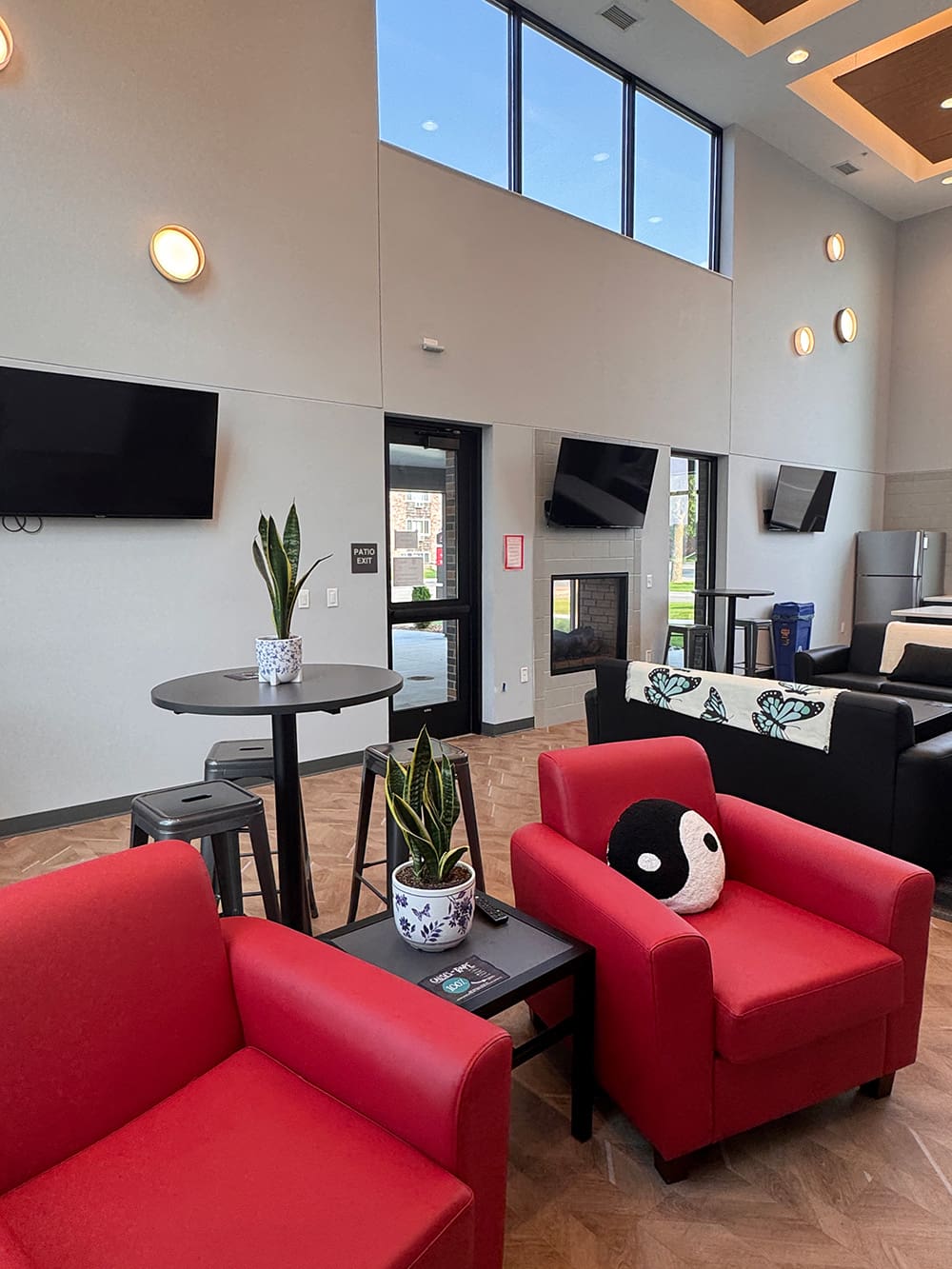 A modern lounge in ATCC Foundation Hall features red armchairs, a small table with potted plants, wall-mounted TVs, large windows, and a high ceiling—bright natural light floods this inviting student housing space.