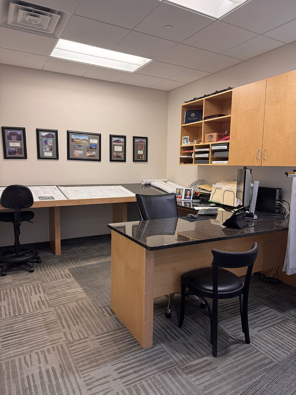 Modern office for innovative builders, featuring a wooden desk, black chair, desktop computer, shelves, framed photos on the wall, and a table with large paper plans. The room has gray carpet and bright overhead lighting.