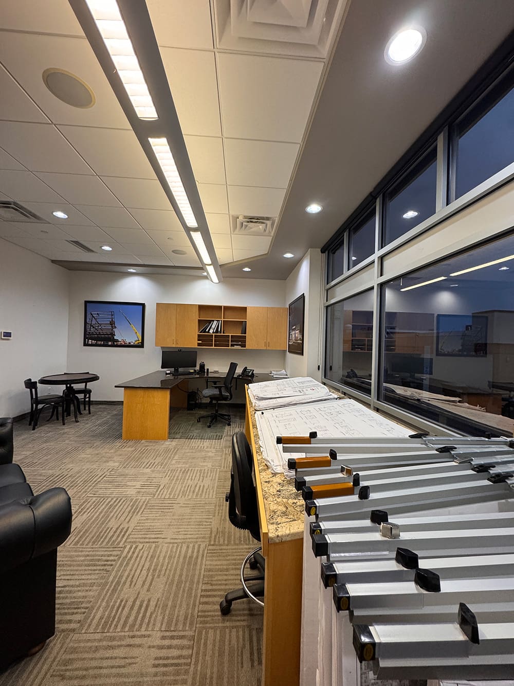 A modern office designed for Innovative Builders features beige carpet, wooden furniture, a desk with a computer, architectural plans on a counter, a black chair, lounge seating, and large windows reflecting the room and cityscape at dusk.