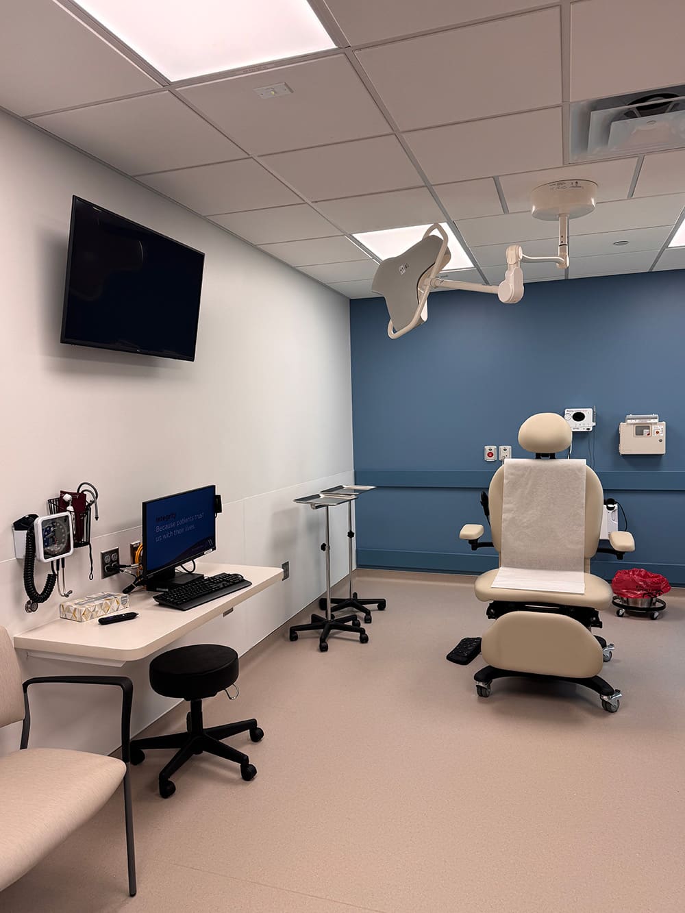A modern medical examination room at Alomere Health features an exam chair, computer workstation, dermatology instruments, overhead exam light, wall-mounted TV, and blue and white walls—ideal for patient care and smooth relocation.