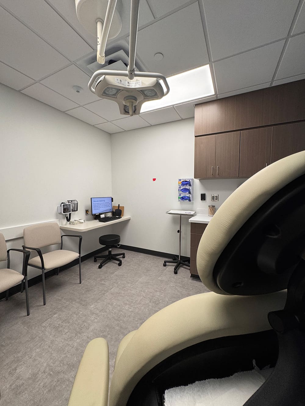 A modern medical exam room at Alomere Health with beige chairs, a black stool, a corner desk with computer, dermatology equipment on the wall, cabinets, ceiling light fixture, and part of an exam chair in the foreground.