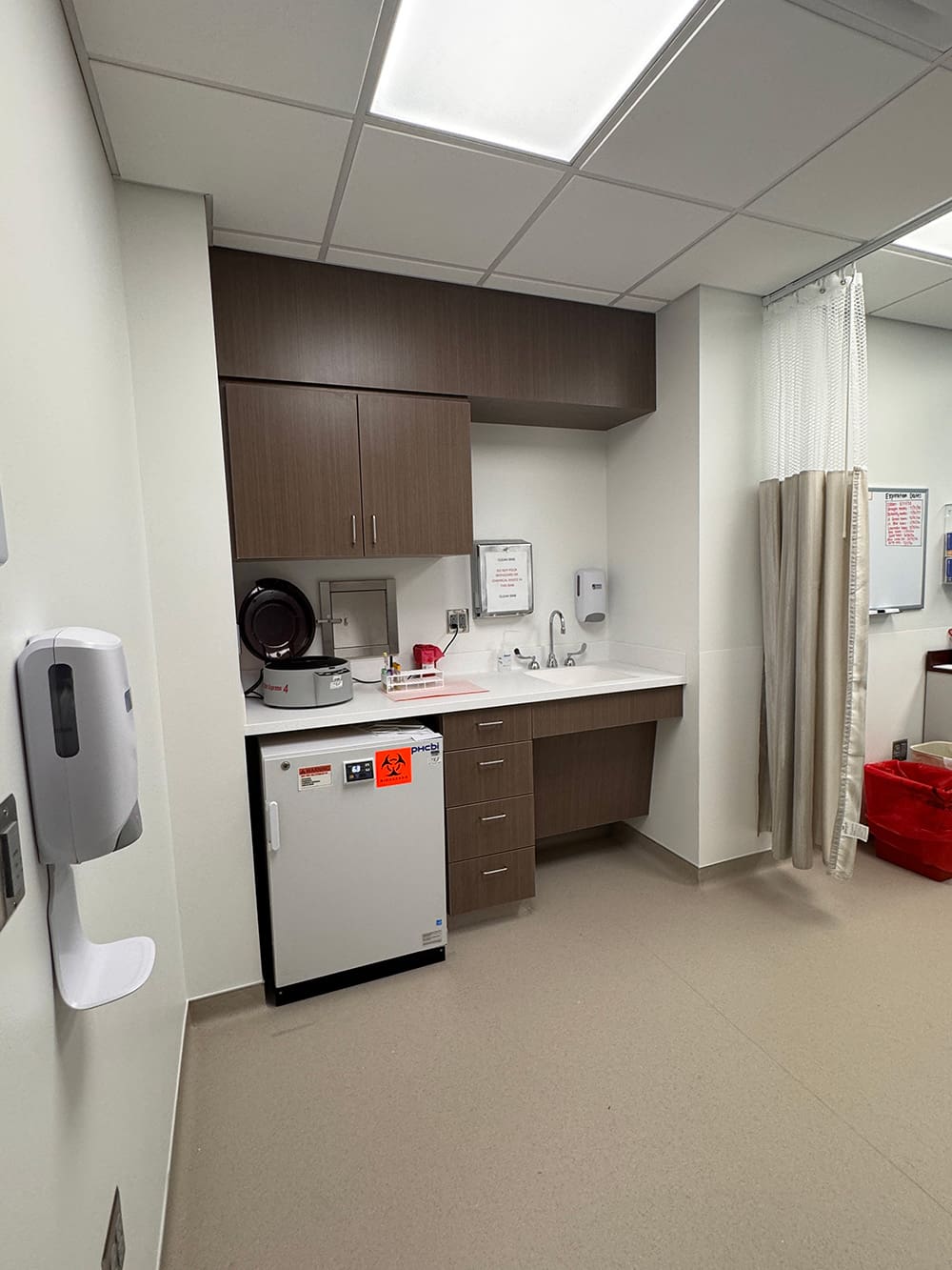 A medical room sink area at Alomere Health features brown cabinets, a white mini fridge with biohazard labels, a soap dispenser, countertop with sink, and medical equipment. A privacy curtain and part of a red bin are visible—ideal for dermatology care.