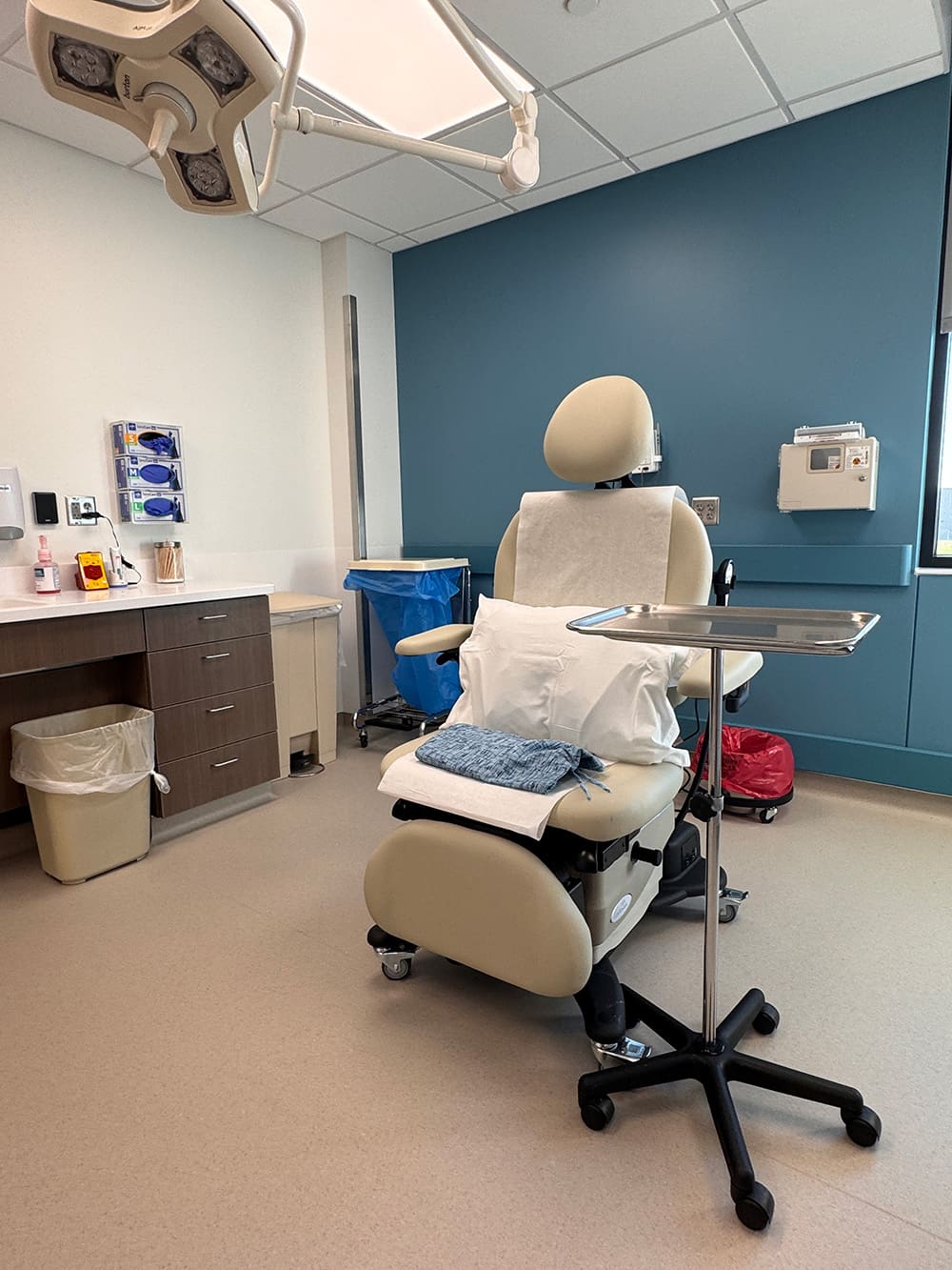 A modern dermatology examination room at Alomere Health features a reclining beige patient chair, a small tray table, folded towels, medical equipment on the wall, a blue accent wall, and overhead exam lights.
