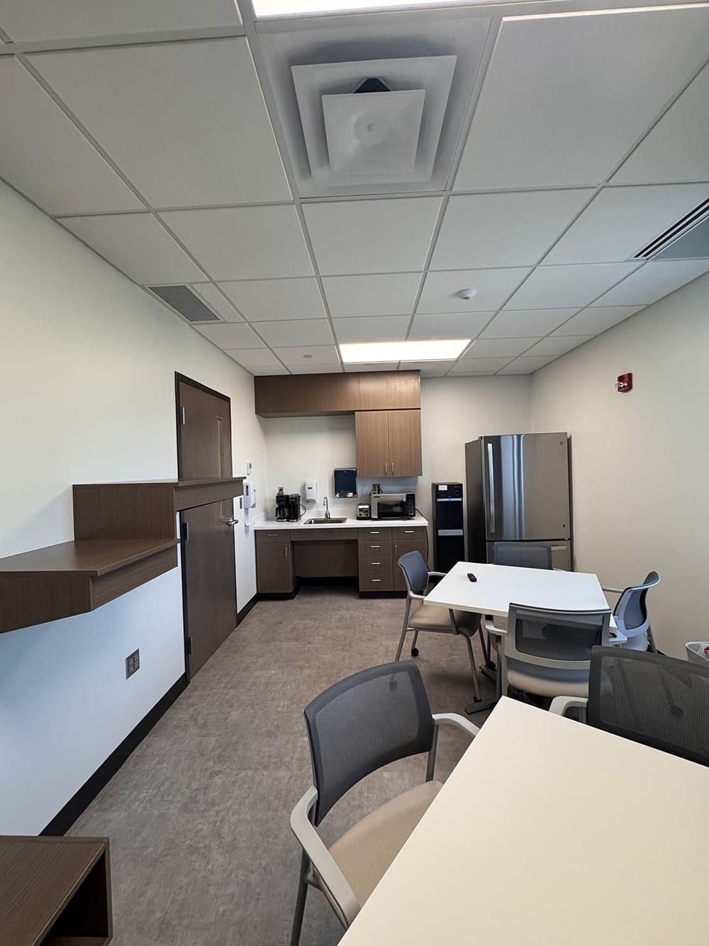 A break room at Alomere Health with tables and chairs, a kitchenette area with a microwave, coffee maker, and stainless steel refrigerator. The room features white walls, a drop ceiling, and gray flooring—ideal for staff in Dermatology or during relocation.