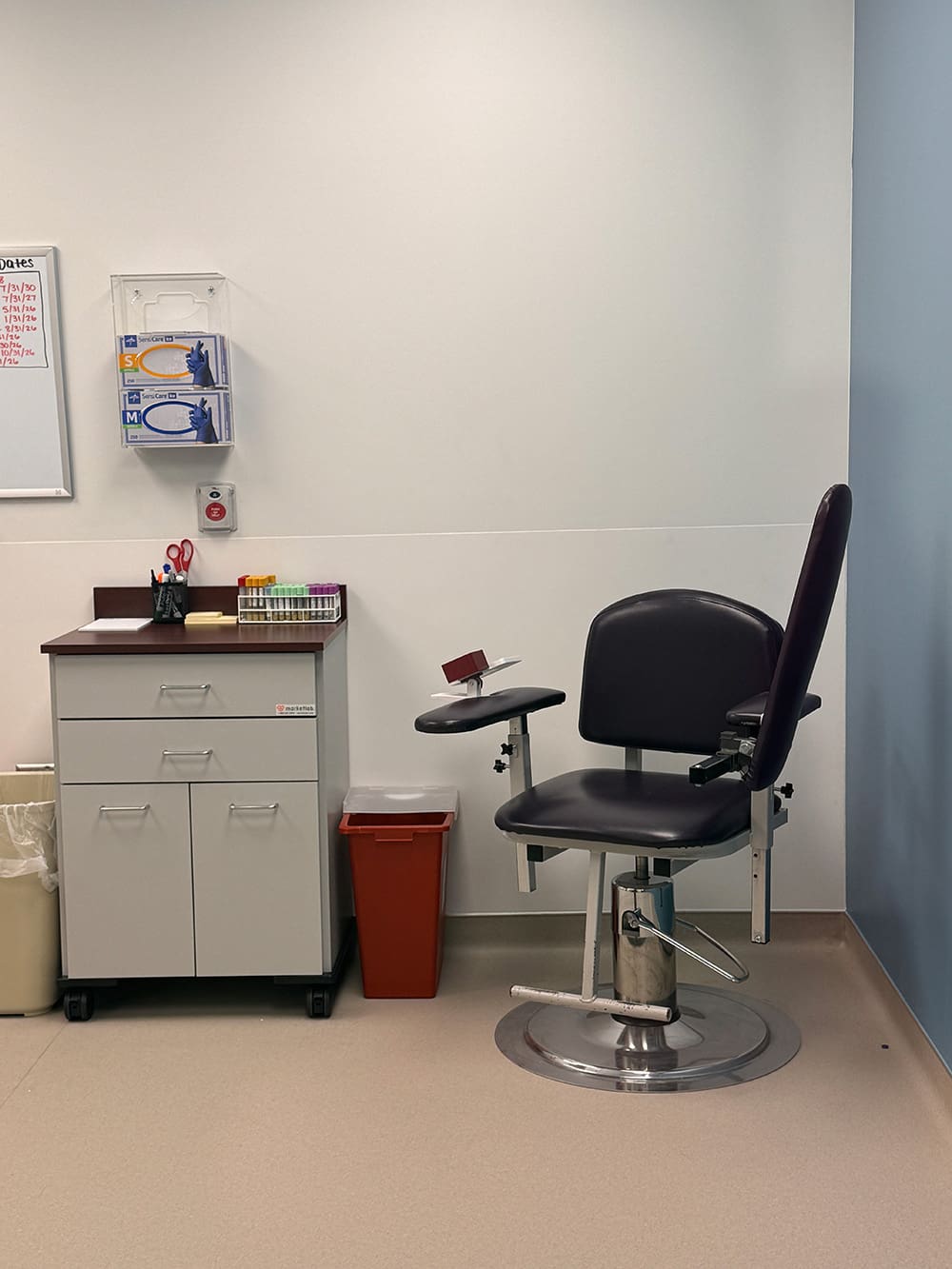A medical exam room at Alomere Health features a black phlebotomy chair, a cabinet with medical supplies, a red sharps container, test tubes, and a trash bin against a white and light blue wall—ideal for dermatology appointments or relocation needs.