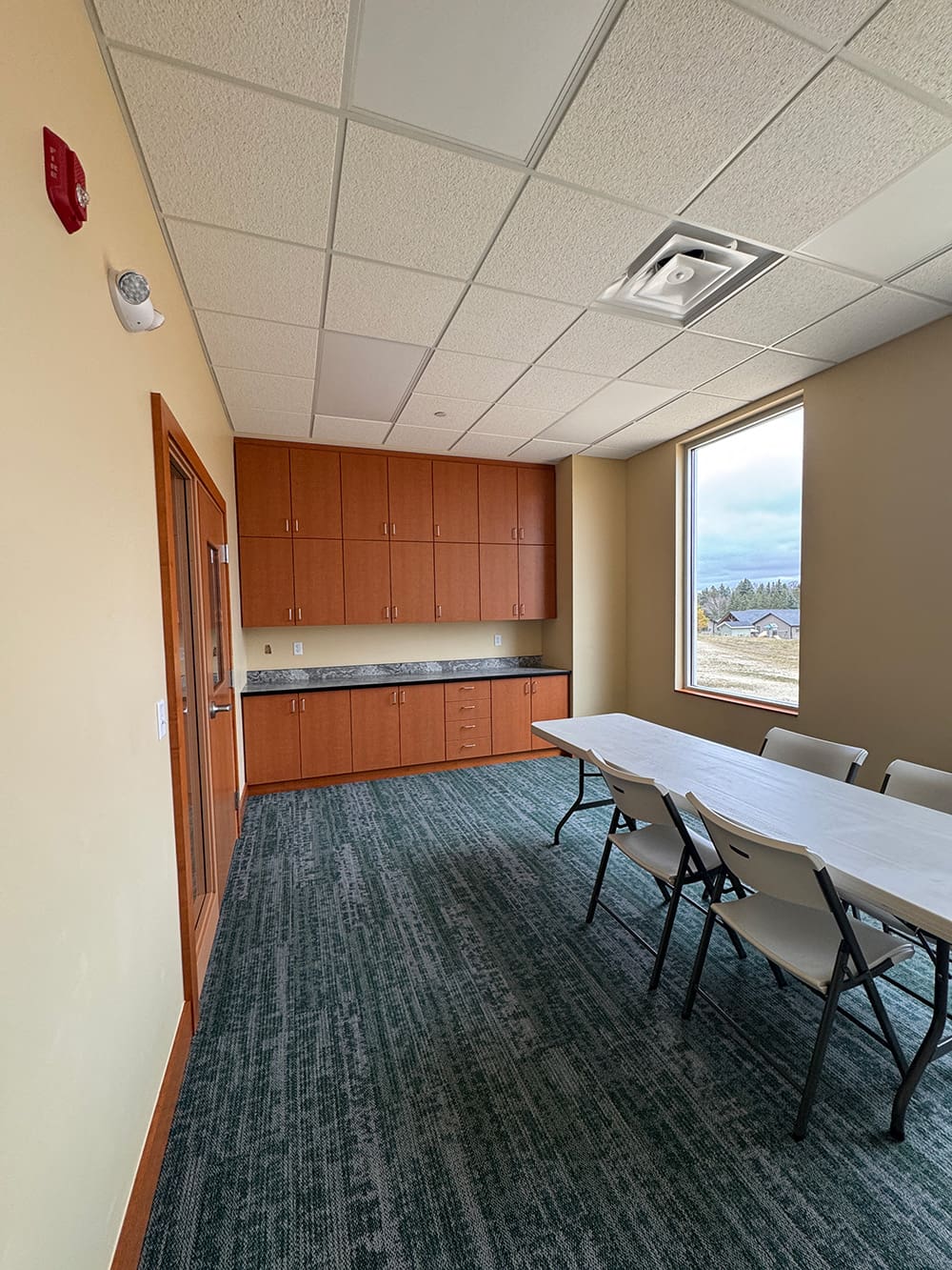 A small meeting room at Living Word Lutheran Church features a long white table, several folding chairs, built-in wooden cabinets along the back wall, carpeted flooring, and a large window overlooking trees and a cloudy sky.