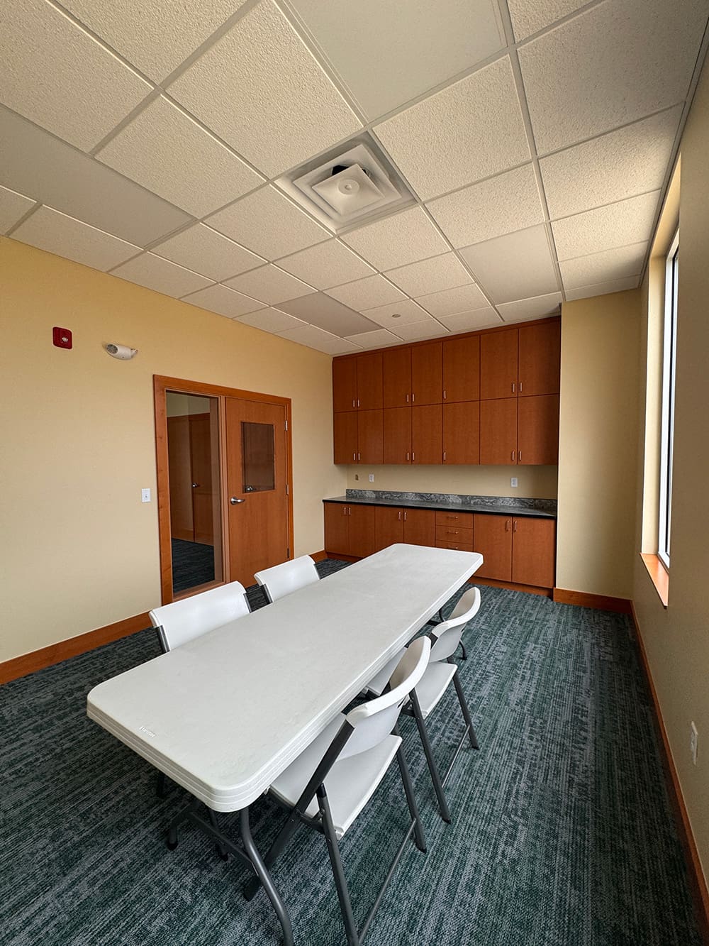 A small conference room at Living Word Lutheran Church features a white table, six white folding chairs, wooden cabinets along the back wall, a carpeted floor, beige walls, and a window letting in natural light.