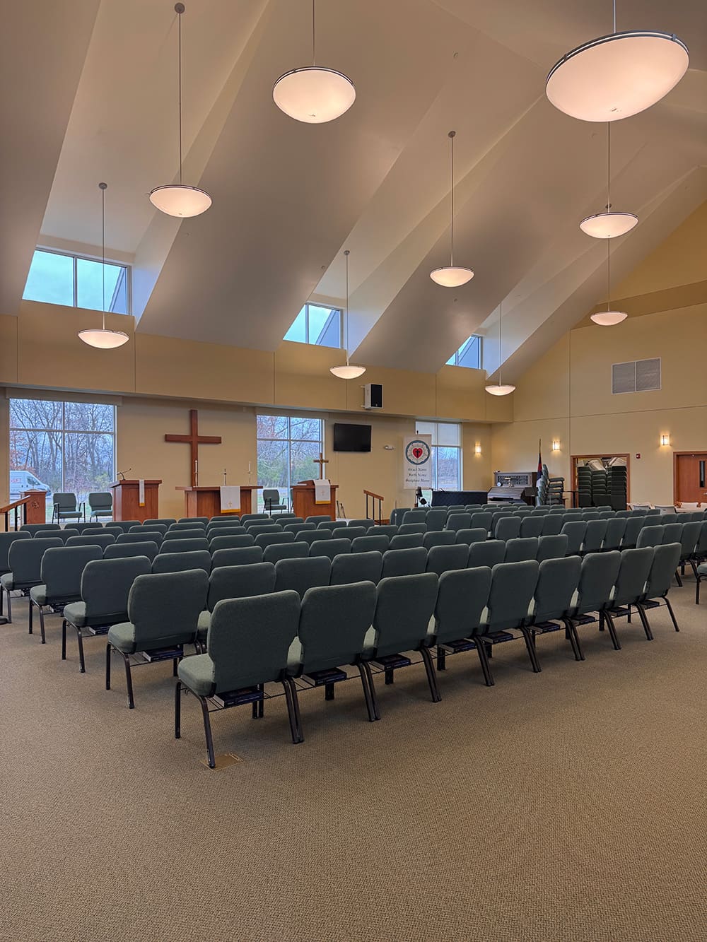 A modern sanctuary at Living Word Lutheran Church features rows of empty green chairs facing a stage with a wooden cross, pulpit, and musical instruments. Large windows let in natural light while round ceiling lights hang overhead.