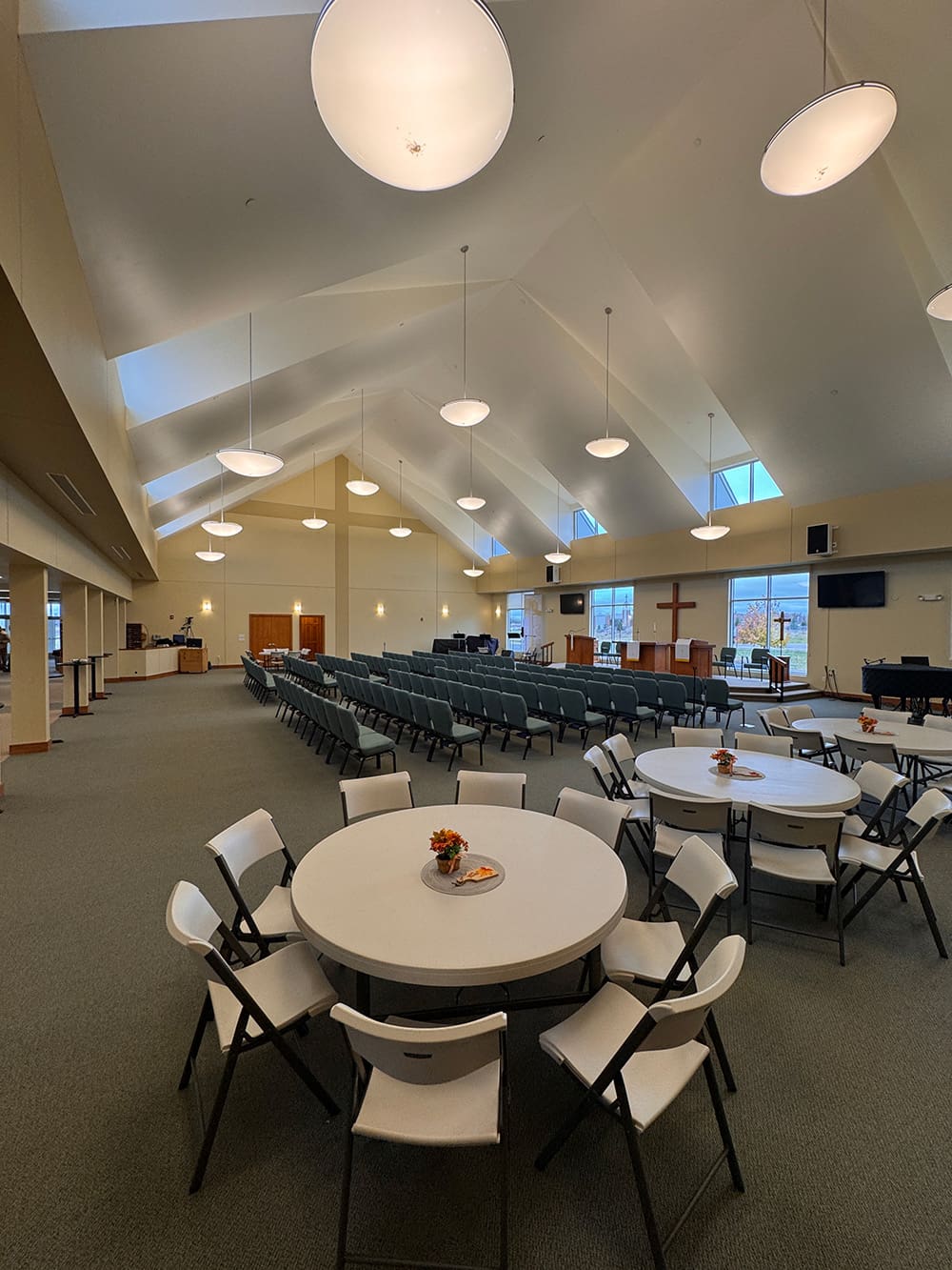 A spacious, well-lit room at Living Word Lutheran Church features high ceilings, round tables with white chairs in the foreground, and rows of chairs facing a stage with a cross and musical instruments in the background.