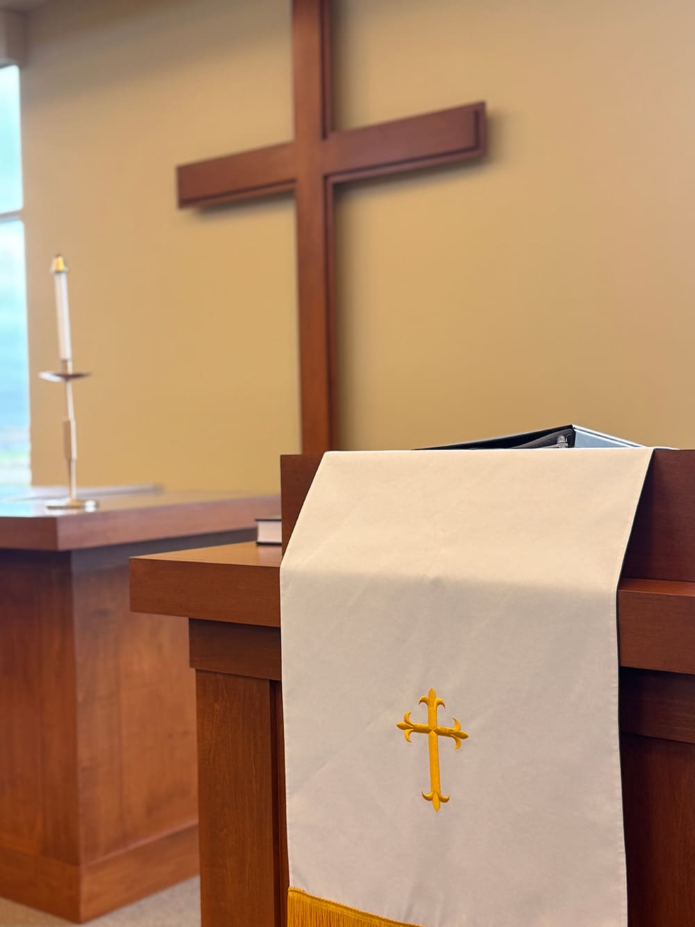 A wooden church pulpit draped with a white cloth featuring a gold cross stands in the foreground at Living Word Lutheran Church, with a large wooden cross and a candle on an altar visible in the background.