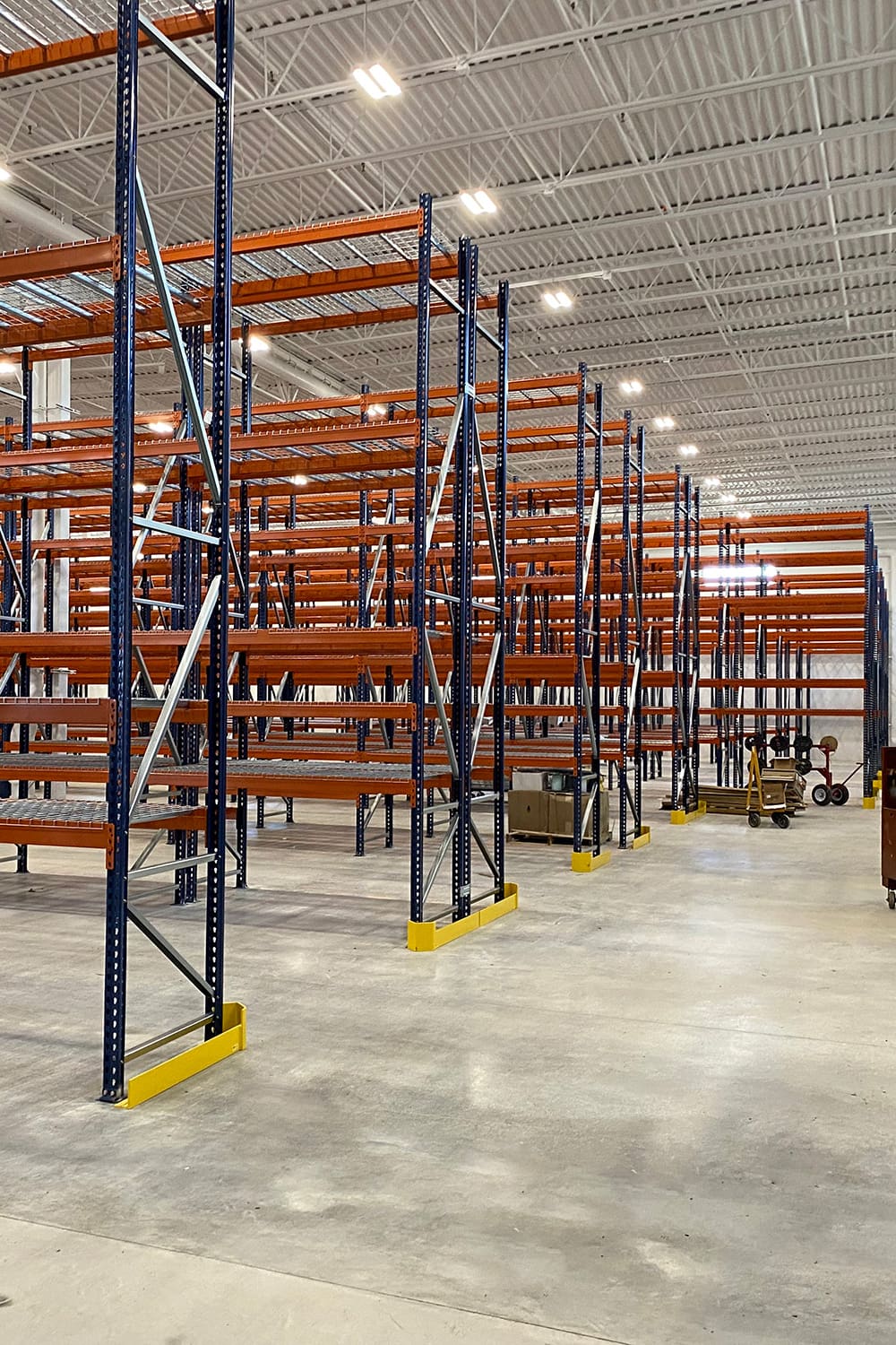 Rows of empty industrial warehouse shelving units from American Solutions for Business stand on a concrete floor under bright ceiling lights, with some equipment and boxes visible in the background.